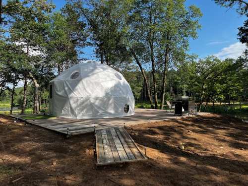 A white geodesic dome on a wooden deck surrounded by trees and blue sky. A grill is visible nearby.