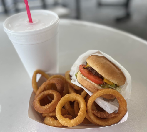 A burger with lettuce and tomato next to a serving of onion rings and a drink with a pink straw on a table.