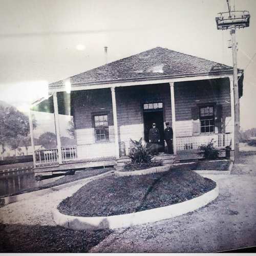 Historic black-and-white photo of a small wooden building with two men standing at the entrance.