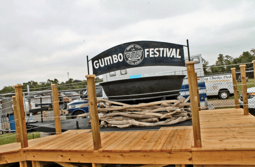 Sign for the Gumbo Festival displayed on a wooden platform, with a large pot and rustic decor in the background.