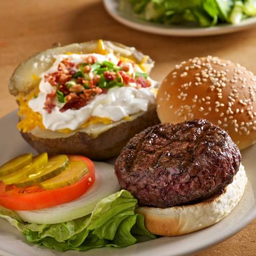 A plate with a grilled burger patty, a sesame seed bun, a baked potato topped with cheese and bacon, and fresh vegetables.