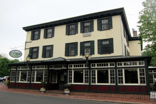 Historic building with a yellow exterior, green shutters, and a welcoming entrance, featuring a sign and outdoor seating.