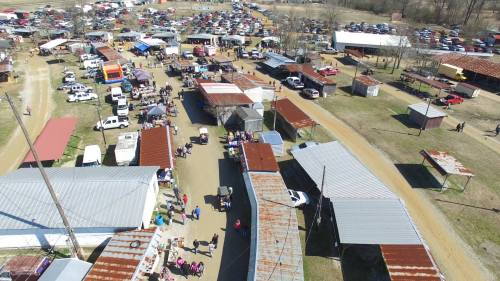 Aerial view of a bustling outdoor market with vendors, cars, and visitors among rustic buildings and tents.