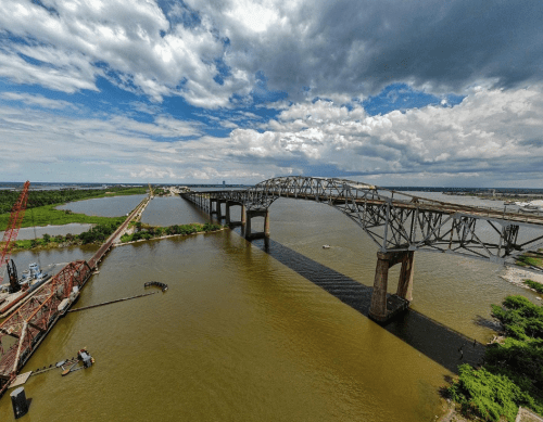 Aerial view of a large bridge spanning a river, with clouds and greenery surrounding the waterway.
