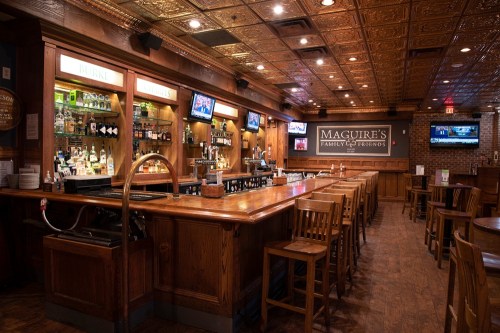 A warm, wooden bar interior with high stools, shelves of liquor, and a cozy atmosphere at Maguire's Family & Friends.