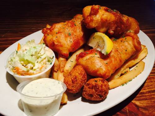 A plate of crispy fish fillets with fries, coleslaw, a lemon wedge, and hush puppies, garnished with herbs.