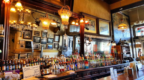 A vintage bar with a wide selection of colorful bottles, ornate lighting, and framed photos on the walls.