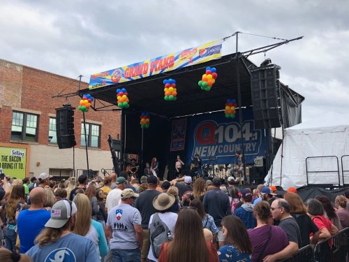 A crowd gathers in front of a large stage with colorful balloons, enjoying a live music performance at an outdoor event.