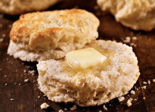 Freshly baked biscuit with a pat of butter melting on top, resting on a wooden surface. Crumbly texture visible.
