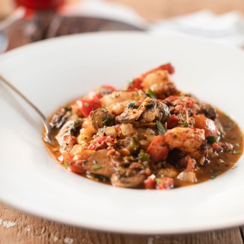 A bowl of shrimp and vegetable stew, garnished with herbs, served with a spoon on a wooden table.