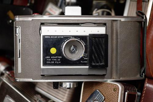 Close-up of a vintage Polaroid camera with a J66 adapter, surrounded by other cameras and accessories.