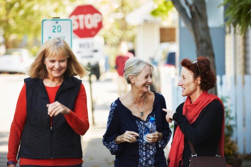 Three women walking together, smiling and chatting on a sunny street, with a stop sign in the background.