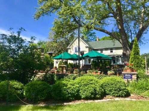 A charming house with green umbrellas over a patio, surrounded by lush greenery and colorful flowers under a clear blue sky.