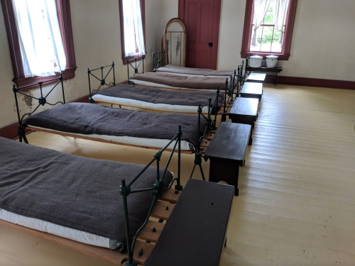 A room with several metal-framed beds lined up, featuring wooden side tables and large windows with white curtains.