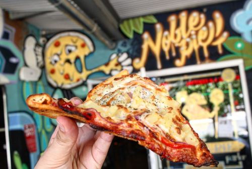 A hand holding a slice of pizza in front of a colorful mural featuring a pizza character.