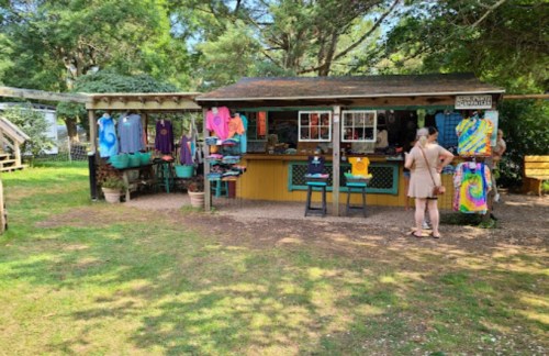A colorful outdoor shop with tie-dye clothing displayed, surrounded by trees and a visitor browsing the items.