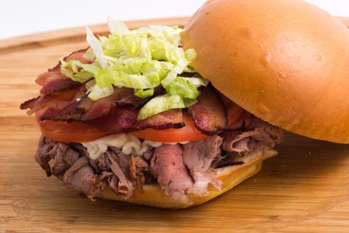 A hearty sandwich with roast beef, bacon, lettuce, tomato, and mayonnaise on a soft bun, served on a wooden board.