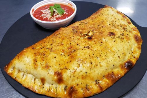 A golden-brown calzone on a black plate, served with a side of marinara sauce topped with cheese and herbs.