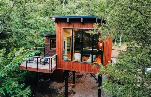 A modern treehouse with large windows, surrounded by lush greenery, featuring a deck and elevated design.