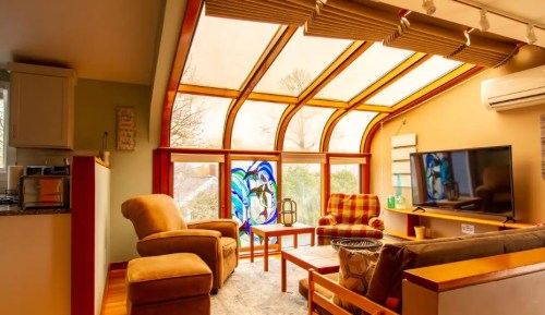Cozy living room with large windows, colorful stained glass, comfortable seating, and a TV. Bright and inviting atmosphere.
