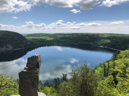 A serene lake surrounded by lush greenery and hills, reflecting clouds in the clear blue sky. A rock formation in the foreground.