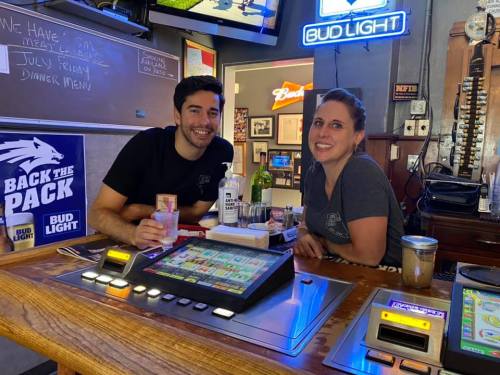 Two smiling bartenders pose behind a bar with drinks and a gaming machine, in a lively pub setting.