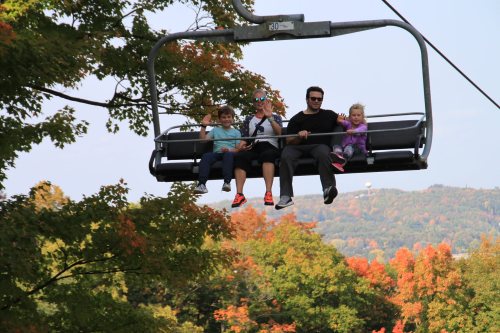A family of four rides a chairlift, smiling against a backdrop of colorful autumn trees.