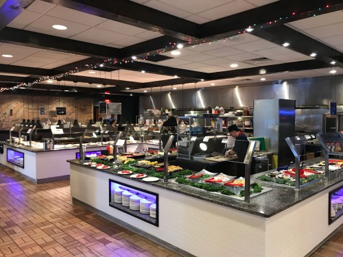 A spacious restaurant buffet with various food stations, colorful decorations, and staff preparing dishes in the kitchen.