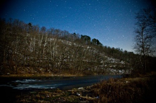 A serene river flows through a wooded landscape under a starry night sky, with a hint of snow on the hillside.