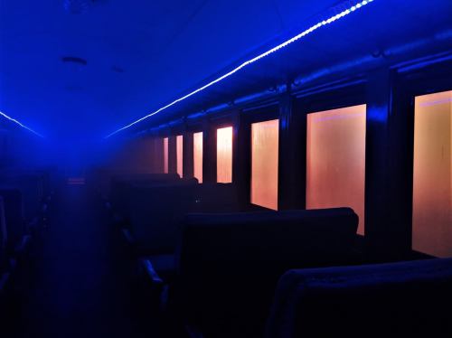 A dimly lit train carriage with blue lighting and fog, featuring rows of empty seats and glowing windows.