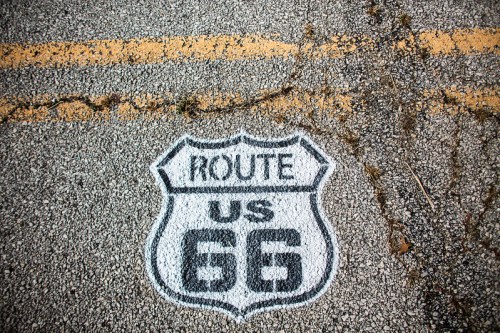 A weathered US Route 66 sign painted on cracked asphalt, surrounded by faded yellow lane markings.
