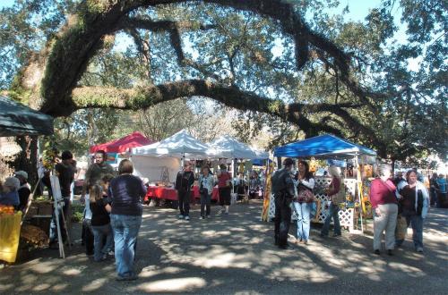 A bustling outdoor market with colorful tents, people browsing, and large trees providing shade.