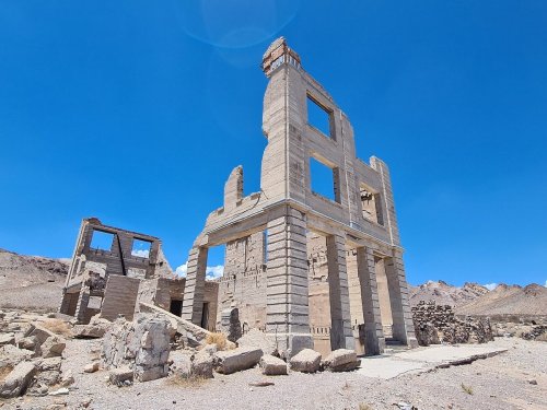 Ruins of a concrete building against a clear blue sky, surrounded by rocky terrain and distant mountains.