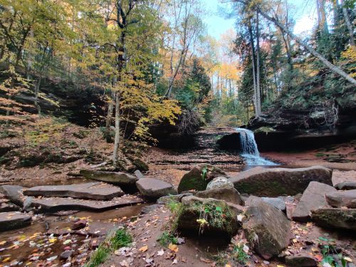 A serene waterfall cascades over rocks in a forest, surrounded by autumn foliage and vibrant trees.