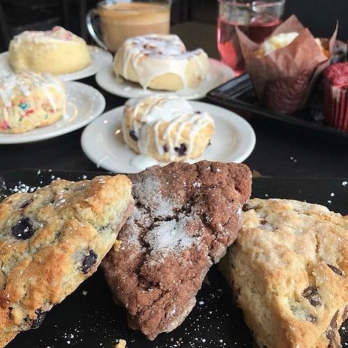 A variety of baked goods including scones, cinnamon rolls, and muffins on a table with drinks in the background.