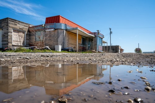 Abandoned building with boarded windows, red roof, and reflection in a puddle on a gravel road under a clear blue sky.
