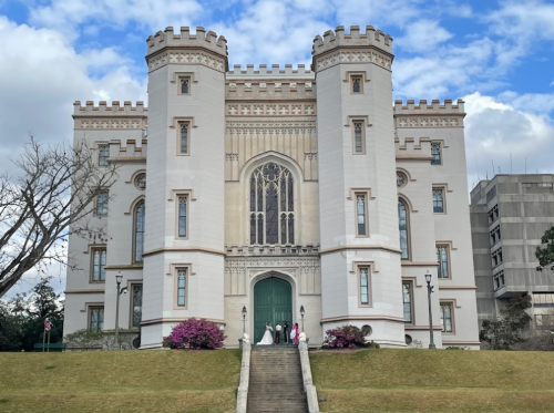 A grand castle-like building with towers, surrounded by greenery and flowers, under a cloudy sky.
