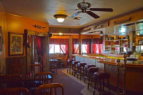 Cozy bar interior with wooden stools, a polished counter, and warm orange walls, featuring red curtains and rustic decor.