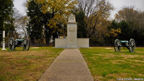 A memorial with a statue and two cannons on either side, surrounded by trees and grass.