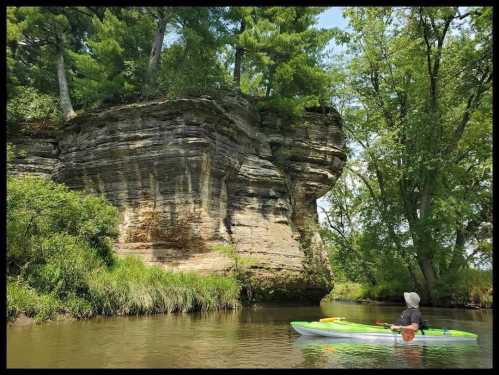 A person in a kayak paddles near a rocky cliff surrounded by lush greenery and trees.