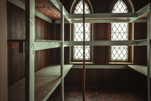 An empty room with wooden walls, shelves, and two large stained glass windows. A rope hangs from the ceiling.