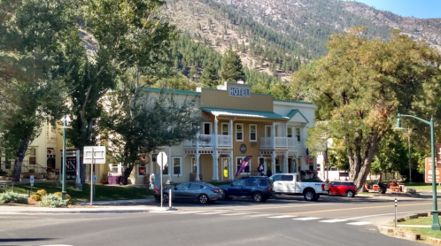 A quaint hotel building surrounded by trees and mountains, with cars parked in front and a clear blue sky.