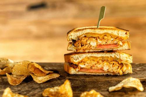 A stacked sandwich with chicken, cheese, and tomato, served with crispy chips on a wooden surface.