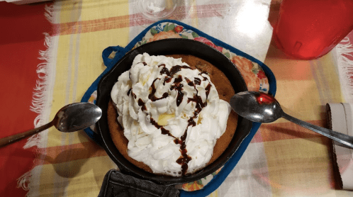 A skillet dessert topped with whipped cream and chocolate drizzle, surrounded by two spoons on a colorful tablecloth.