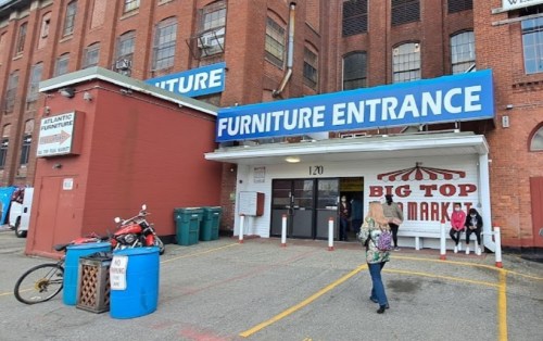 A red brick building with a large sign reading "Furniture Entrance" and a market sign, with people and a bike nearby.