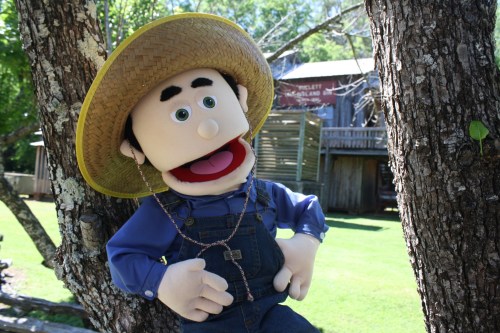 A cheerful puppet wearing a straw hat and overalls leans against a tree, set in a rustic outdoor environment.