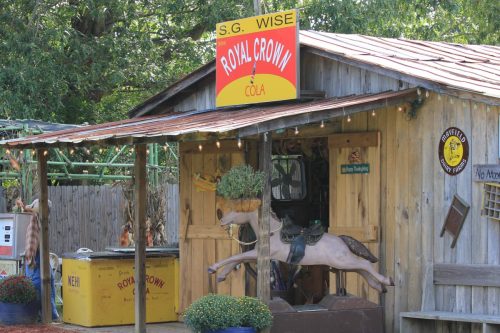 A rustic wooden shed with a red roof, featuring a vintage Royal Crown Cola sign and a horse carousel outside.