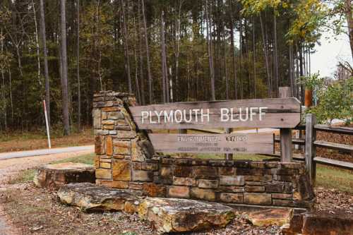 Sign for Plymouth Bluff Environmental Center, surrounded by trees, with a stone base and a gravel road nearby.