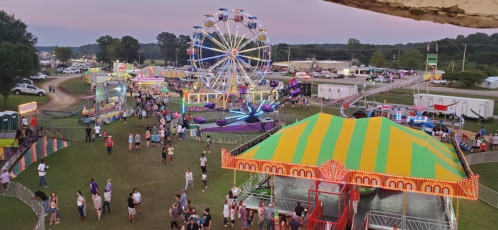 A vibrant carnival scene with a Ferris wheel, colorful tents, and crowds enjoying various attractions at dusk.