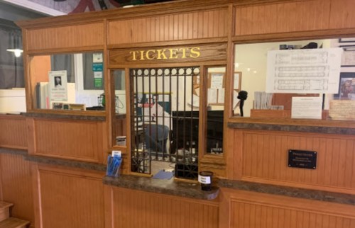 A wooden ticket counter with a metal grate, featuring a sign that reads "TICKETS" and various informational papers.